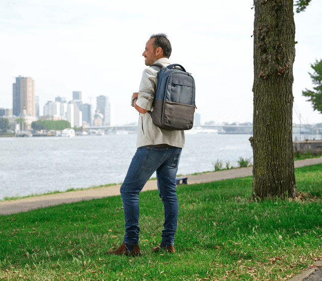 Hombre caminando al aire libre junto al río con la mochila para notebook Lisboa 16 pulgadas de Trust en color azul, con una ciudad moderna de fondo.