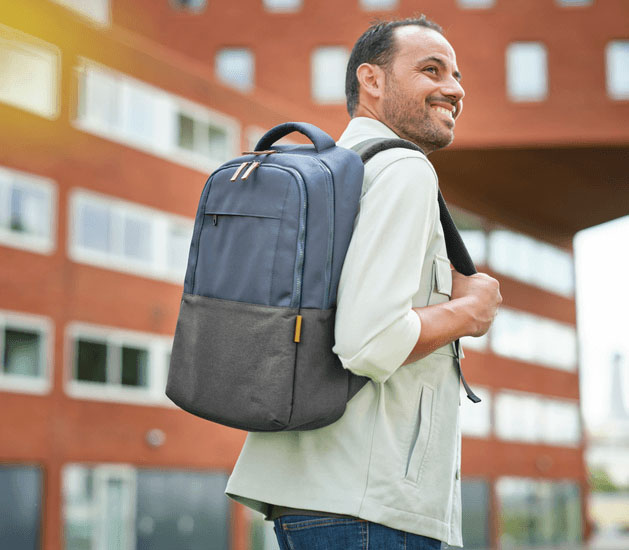Hombre sonriente con la mochila para notebook Trust Lisboa 16 pulgadas en color azul, caminando frente a un edificio moderno de ladrillo.