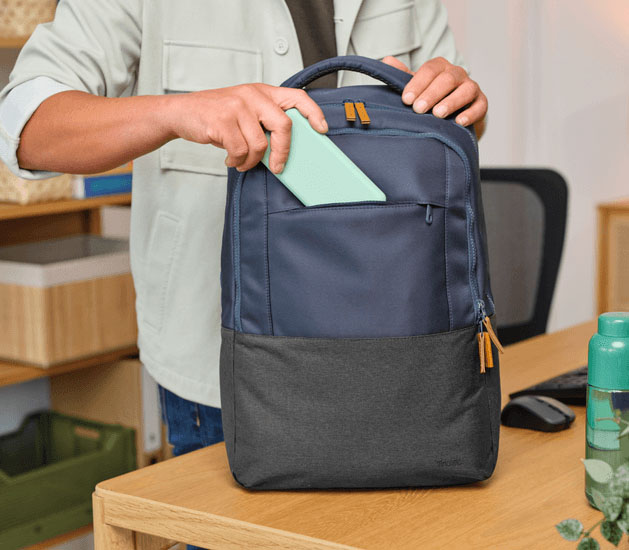 Persona guardando un objeto en el bolsillo frontal de la mochila para notebook Trust Lisboa 16 pulgadas en color azul, apoyada sobre un escritorio en un entorno de oficina moderna.