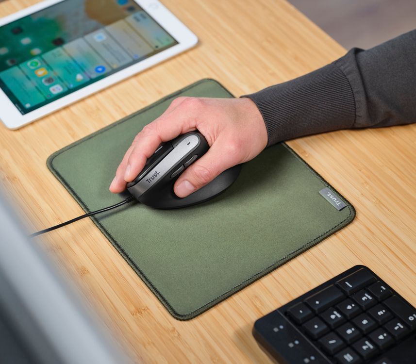 Una mano usando un mouse ergonómico Trust Bayo II con cable sobre una alfombrilla verde en un escritorio de madera clara. En la esquina superior izquierda se ve una tableta y en la esquina inferior derecha, un teclado negro.