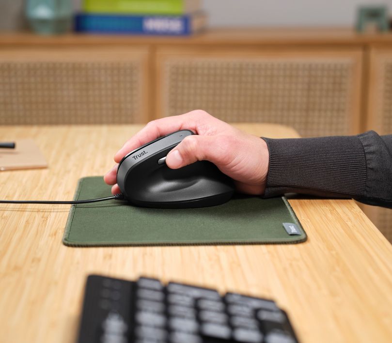 Una mano usando un Mouse ergonómico Trust Bayo II - Negro sobre una alfombrilla verde en un escritorio de madera. Se puede ver un teclado desenfocado y algunos libros.