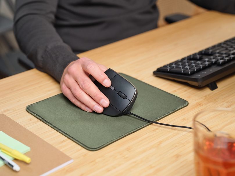 Una mano usando un Mouse ergonómico Trust Bayo II - Negro con cable sobre una alfombrilla verde, junto a un teclado negro en un escritorio de madera clara. También hay una bebida en un vaso y algunos artículos de papelería, como un cuaderno y bolígrafos.