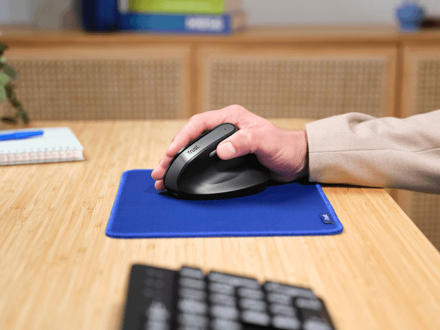 Hombre trabajando en una computadora en su escritorio, usando el mouse ergonómico inalámbrico Trust Bayo II negro sobre un pad para mouse azul.