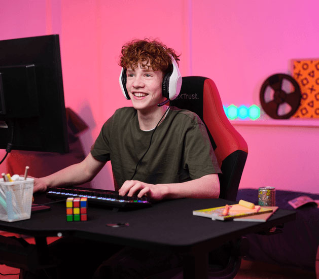 Adolescente con Auriculares Gaming Livianos Trust GXT 415W Zirox de color blanco, jugando en una PC, sentado en una silla ergonómica frente a un escritorio con teclado RGB y útiles, en una habitación iluminada con luces LED rosas.