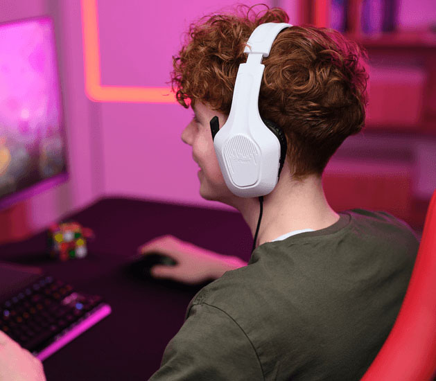 Adolescente con Auriculares Gaming Livianos Trust GXT 415W Zirox de color blanco, jugando en una PC, sonriendo mientras usa el mouse, frente a un monitor con iluminación LED rosa y teclado retroiluminado.