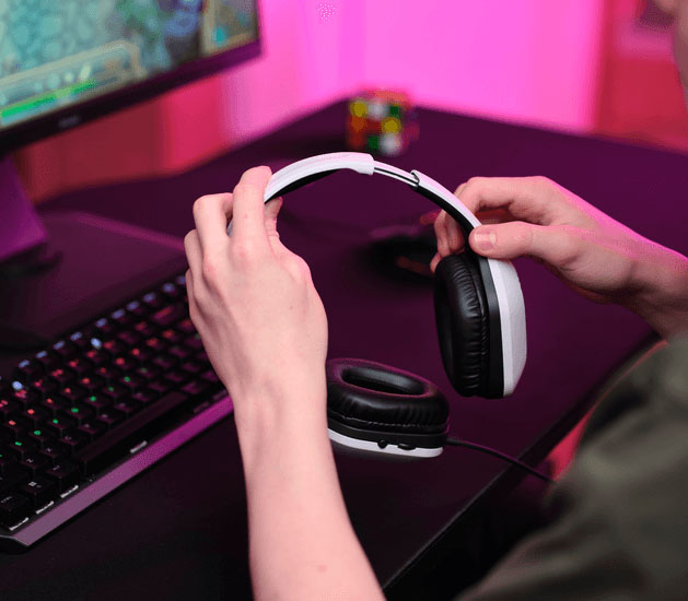 Primer plano de manos ajustando unos Auriculares Gaming Livianos Trust GXT 415W Zirox de color blanco, frente a un monitor con videojuego, teclado mecánico retroiluminado y luces LED rosas de fondo.