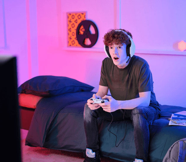 Adolescente sentado en la cama jugando con un joystick y Auriculares Gaming Livianos Trust GXT 415W Zirox de color blanco, disfrutando de un videojuego frente a una pantalla, en una habitación con luces LED en tonos rosa y azul.