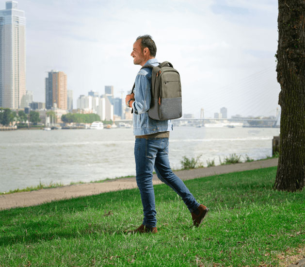 Hombre caminando al aire libre junto al río con la mochila para notebook Lisboa 16 pulgadas de Trust en color verde, con una ciudad moderna de fondo.