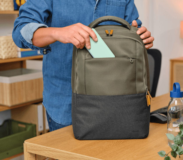 Persona guardando un objeto en el bolsillo frontal de la mochila para notebook Trust Lisboa 16 pulgadas en color verde, apoyada sobre un escritorio en un entorno de oficina moderna.