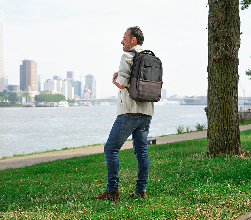 Una persona de pie en un parque junto a un árbol, con una mochila Trust Lisboa negra en la espalda, mirando hacia un río con una ciudad y varios edificios altos al fondo.