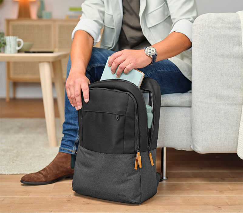 Una persona está sentada en un sofá, guardando una libreta verde en una mochila Trust Lisboa negra con detalles en gris. La mochila está abierta y se encuentra en el suelo, junto a una mesa de madera clara con una taza y otros objetos encima. La persona lleva un reloj en la muñeca y botas marrones.