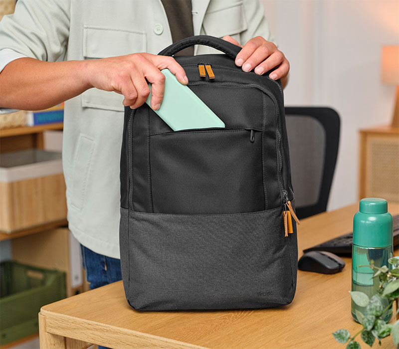 Una persona está guardando una tableta verde en el bolsillo frontal de una mochila Trust Lisboa negra sobre un escritorio de madera. En el escritorio también hay una botella verde, un mouse y un teclado.