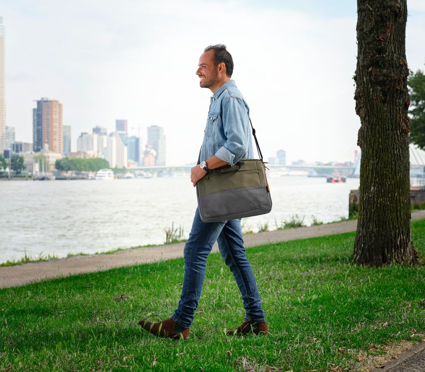 Una persona de pie en un parque junto a un río, con una ciudad moderna y un puente al fondo. Lleva una chaqueta clara, jeans y zapatos marrones, y sostiene un Maletín para Laptop de 16 pulgadas Trust Lisboa de color verde