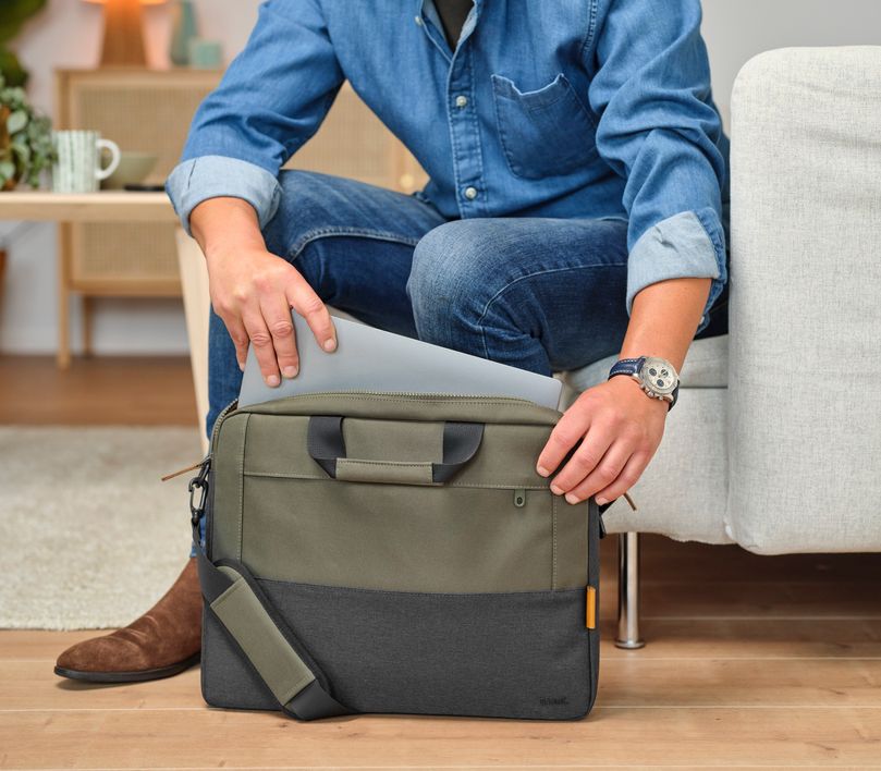 Una persona está sentada en un sofá, sacando una laptop de un Maletín para Laptop de 16 pulgadas Trust Lisboa de color verde. La persona lleva una camisa de jean, jeans y un reloj en la muñeca izquierda. En el fondo, se puede ver una mesa con una taza y algunos objetos decorativos.
