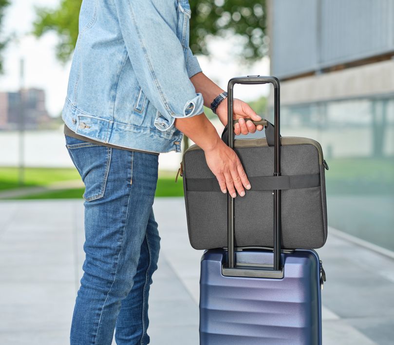 Una persona con una chaqueta clara y jeans está colocando un Maletín para Laptop de 16 pulgadas Trust Lisboa de color verde sobre el asa extendida de una maleta de viaje azul. La escena se desarrolla en un entorno exterior, posiblemente cerca de un edificio moderno y un área verde.