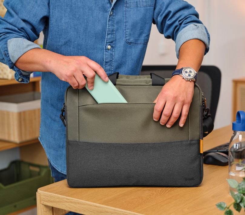 Una persona está guardando un objeto rectangular verde en el bolsillo frontal de un Maletín para Laptop de 16 pulgadas Trust Lisboa de color verde. La persona lleva una chaqueta clara y un reloj plateado en la muñeca izquierda. En la mesa de madera hay una botella verde y algunos objetos más en el fondo.