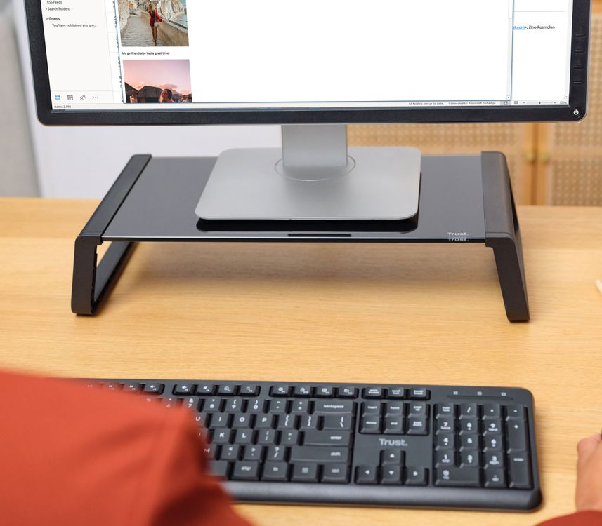 Monitor de computadora sobre un soporte para monitor de cristal templado Trust Monta de color Negro, con teclado en primer plano, sobre un escritorio de madera frente a una persona.