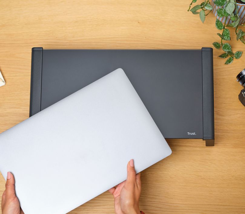 Una persona está colocando una laptop plateada sobre un soporte para monitor de cristal templado Trust Monta de color Negro, sobre un escritorio de madera