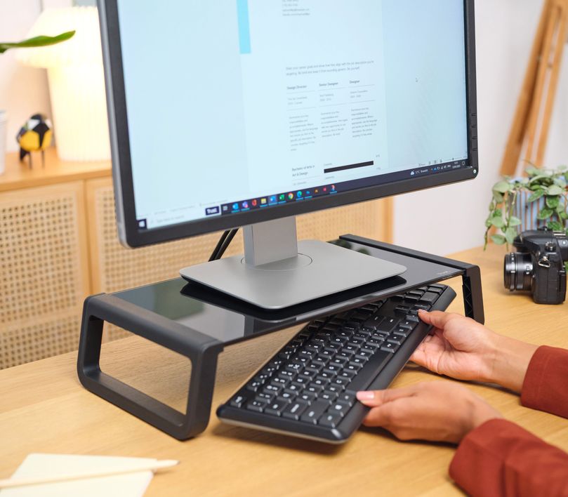 Una persona guardando un teclado debajo de un Soporte para monitor de cristal templado Trust Monta de color Negro, en un escritorio de madera. Hay una planta y una cámara fotográfica al lado del monitor.