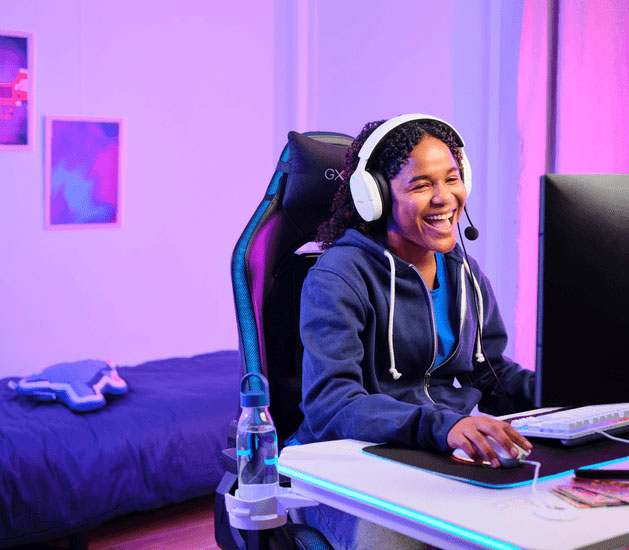 Alguien sentada en una silla gamer con Auriculares Gaming 7.1 USB Trust GXT 490W Fayzo, sonriendo mientras mira un monitor en su dormitorio con iluminación de colores.