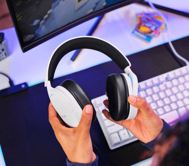 Manos sosteniendo Auriculares Gaming 7.1 USB Trust GXT 490W Fayzo blancos con orejeras negras cerca de un escritorio con teclado y monitor.