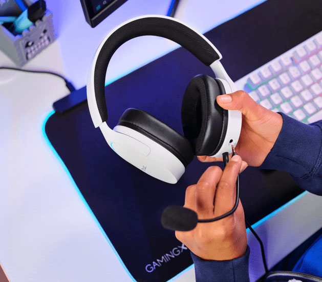 Manos conectando un micrófono desmontable a unos Auriculares Gaming 7.1 USB Trust GXT 490W Fayzo blancos con orejeras negras, cerca de un teclado y mousepad.