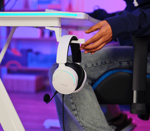 Auriculares Gaming 7.1 USB Trust GXT 490W Fayzo blancos con micrófono colgando de un gancho bajo un escritorio, con una mano cerca, mostrando una opción de almacenamiento.