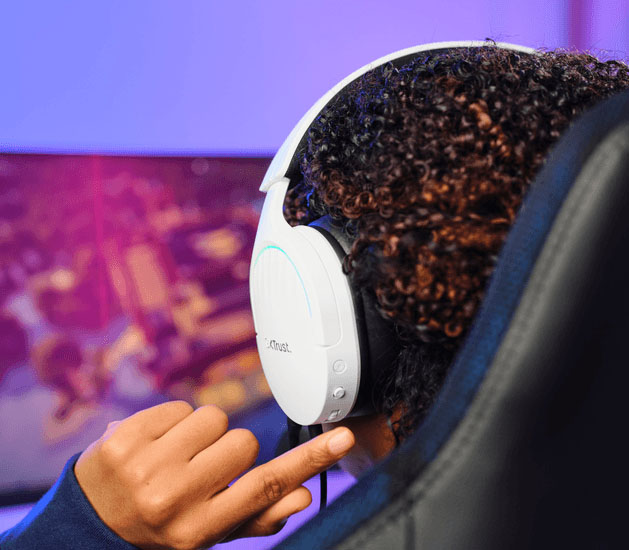 Primer plano de una persona usando Auriculares Gaming 7.1 USB Trust GXT 490W Fayzo blancos con micrófono, mirando un monitor de juego desenfocado.