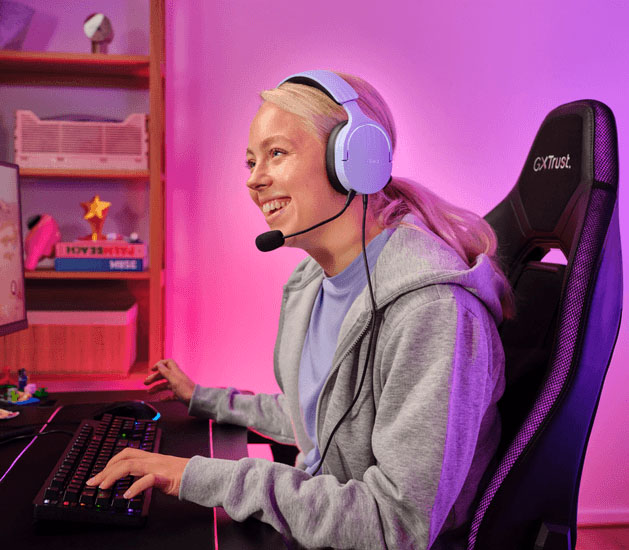 Alguien sentada en una silla gamer con Auriculares Gaming 7.1 USB Trust GXT 490P Fayzo, sonriendo mientras mira un monitor en su dormitorio con iluminación de colores.