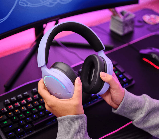 Manos sosteniendo Auriculares Gaming 7.1 USB Trust GXT 490P Fayzo Morados con orejeras negras cerca de un escritorio con teclado y monitor.