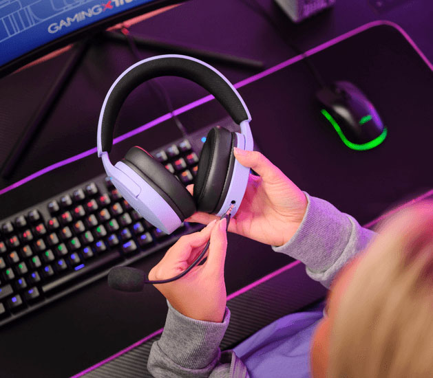 Manos conectando un micrófono desmontable a unos Auriculares Gaming 7.1 USB Trust GXT 490P Fayzo Morados con orejeras negras, cerca de un teclado y mousepad.