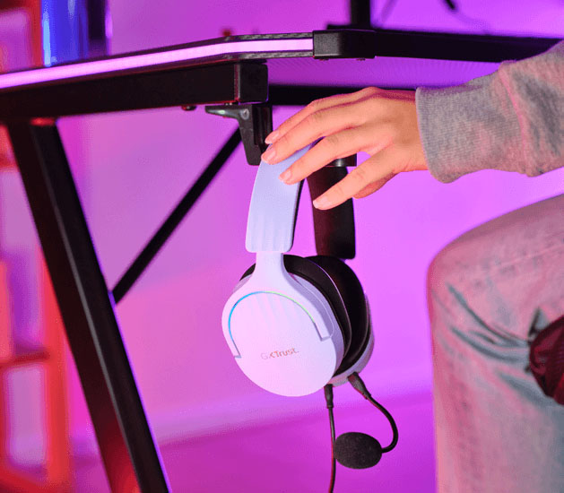 Auriculares Gaming 7.1 USB Trust GXT 490P Fayzo Morados con micrófono colgando de un gancho bajo un escritorio, con una mano cerca, mostrando una opción de almacenamiento.
