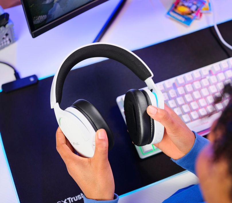 Una persona sosteniendo unos Auriculares Gaming Inalámbricos Trust GXT 491W Fayzo, de color blanco sobre un escritorio con accesorios de gaming.