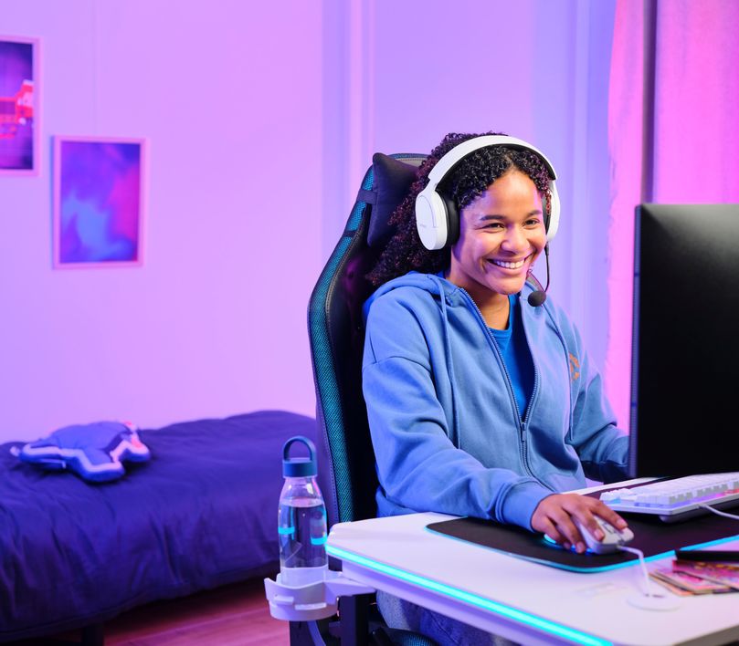 Persona sentada frente a una computadora con Auriculares Gaming Inalámbricos Trust GXT 491W Fayzo, de color blanco, jugando o trabajando en un ambiente iluminado con luces violetas.