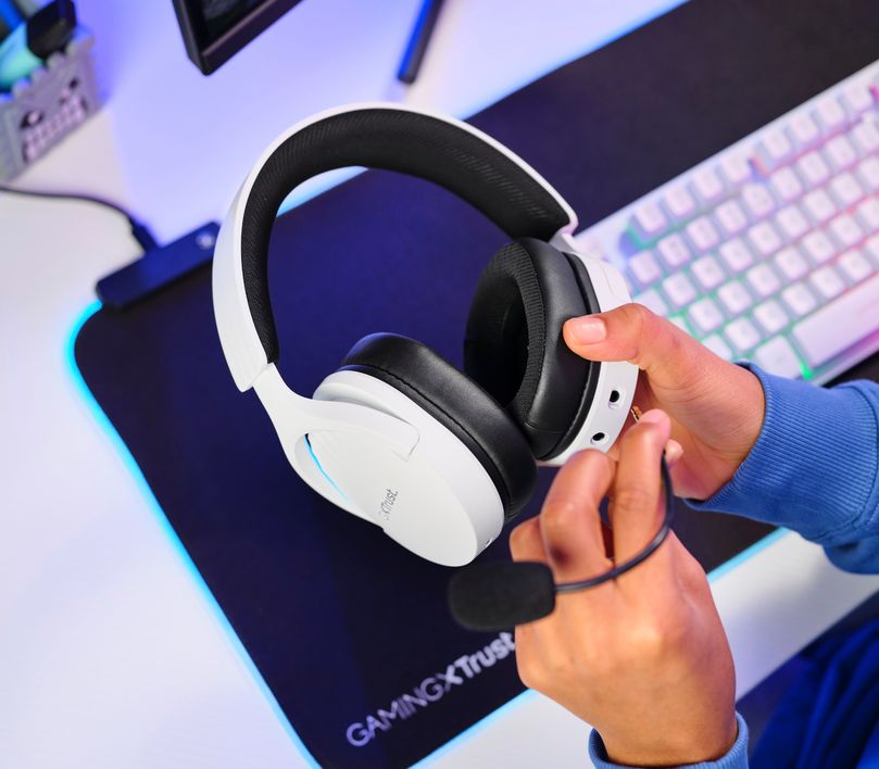 Manos sosteniendo unos Auriculares Gaming Inalámbricos Trust GXT 491W Fayzo, de color blanco sobre un escritorio con teclado y mousepad iluminados por luces LED.
