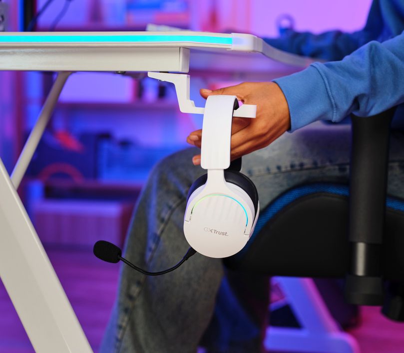 Auriculares Gaming Inalámbricos Trust GXT 491W Fayzo, de color blanco colgando de un soporte para auriculares adherido a una mesa, con una persona al fondo tocandolo, iluminado por luces azules y violetas.