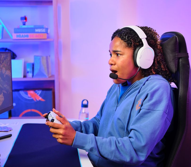 Persona jugando videojuegos con un controlador, usando unos Auriculares Gaming Inalámbricos Trust GXT 491W Fayzo, de color blanco, sentada en una silla de oficina frente a un escritorio con una computadora y monitor iluminados por luces LED azules.
