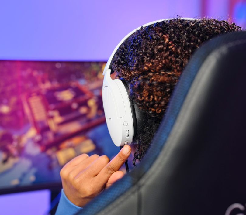Persona con Auriculares Gaming Inalámbricos Trust GXT 491W Fayzo, de color blanco ajustando el volumen en un dial lateral, sentada frente a una computadora con pantalla mostrando gráficos coloridos de un videojuego.