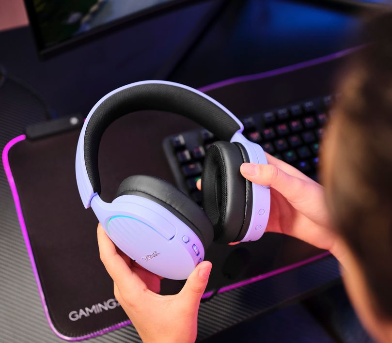 Una persona sosteniendo unos Auriculares Gaming Inalámbricos Trust GXT 491P Fayzo, de color Morado sobre un escritorio con accesorios de gaming.