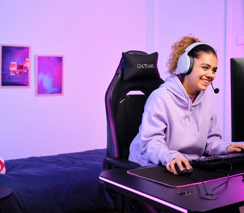 Persona sentada frente a una computadora con Auriculares Gaming Inalámbricos Trust GXT 491P Fayzo, de color Morado, jugando o trabajando en un ambiente iluminado con luces violetas.