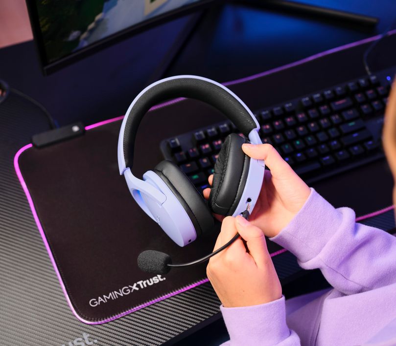 Manos sosteniendo unos Auriculares Gaming Inalámbricos Trust GXT 491P Fayzo, de color Morado sobre un escritorio con teclado y mousepad iluminados por luces LED.