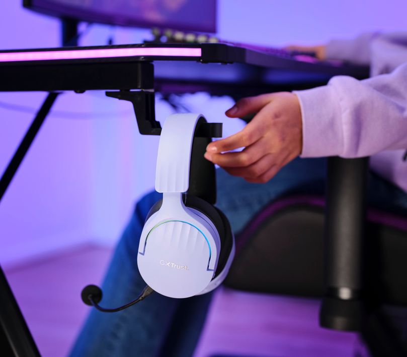 Auriculares Gaming Inalámbricos Trust GXT 491P Fayzo, de color Morado colgando de un soporte para auriculares adherido a una mesa, con una persona al fondo tocandolo, iluminado por luces azules y violetas.