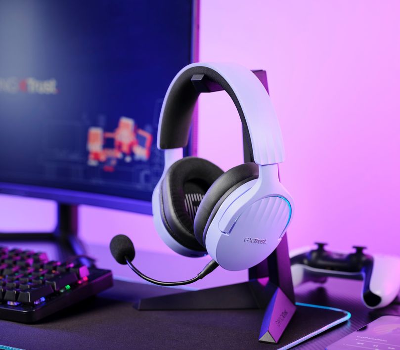 Unos Auriculares Gaming Inalámbricos Trust GXT 491P Fayzo, de color Morado, , apoyados en un soporte, frente a una pantalla de computadora y un teclado en un fondo iluminado con luces violetas y azules.
