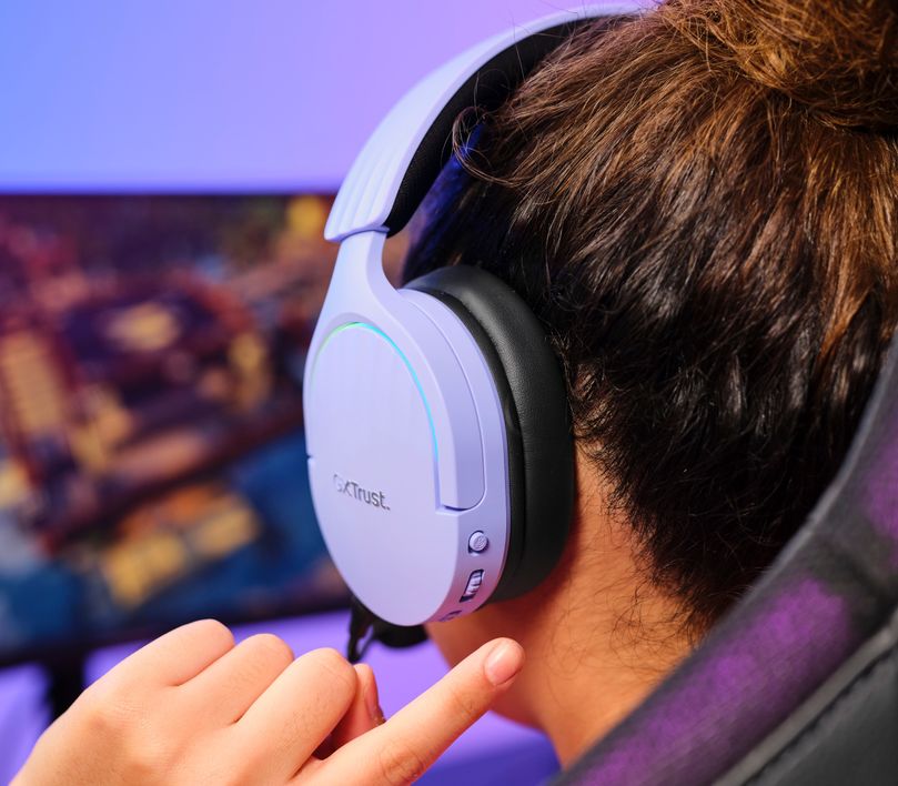 Persona con Auriculares Gaming Inalámbricos Trust GXT 491P Fayzo, de color Morado ajustando el volumen en un dial lateral, sentada frente a una computadora con pantalla mostrando gráficos coloridos de un videojuego.