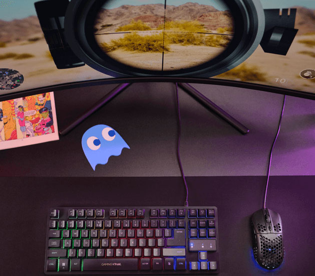 Mouse gaming Trust GXT 928 Helox ultraliviano con iluminación RGB sobre un escritorio gamer junto a un teclado compacto y un monitor curvo