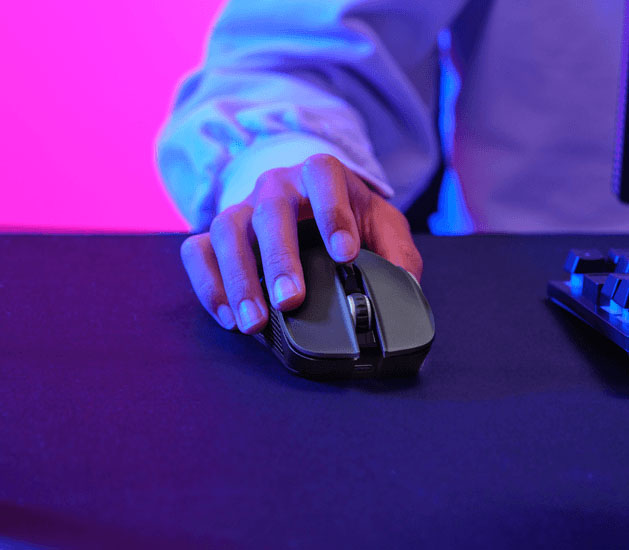 Mano usando un Mouse gaming inalámbrico multidispositivo Trust GXT 931 Ybar sobre una superficie oscura con un fondo iluminado en rosa y azul.