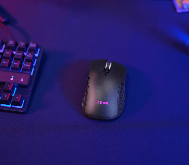 Vista superior lateral de un Mouse gaming inalámbrico multidispositivo Trust GXT 931 Ybar sobre una superficie oscura, con un teclado al lado y fondo iluminado en rosa y azul.