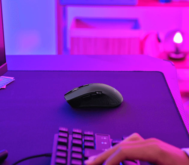 Vista lateral de un Mouse gaming inalámbrico multidispositivo Trust GXT 931 Ybar sobre un mousepad oscuro, con una mano y parte de un teclado en primer plano y fondo iluminado en rosa y azul.