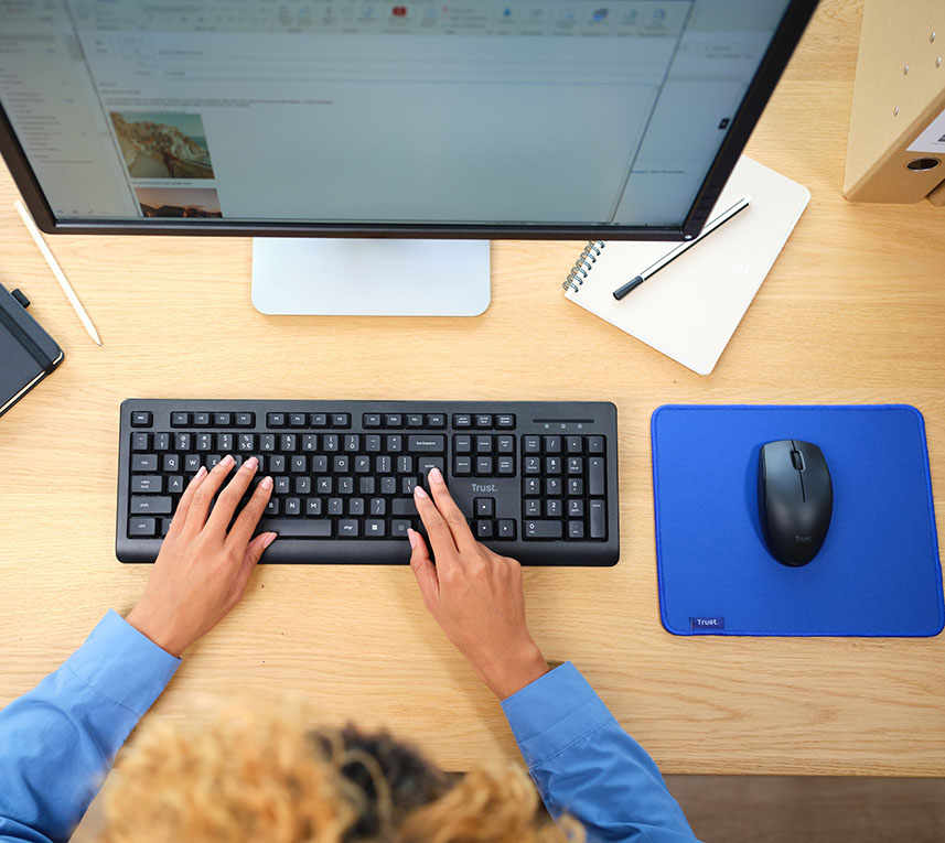 Vista superior de un escritorio con una computadora y accesorios; una persona está utilizando un Combo de Teclado y Mouse inalámbricos Trust Primo, al lado hay un cuaderno con bolígrafo.