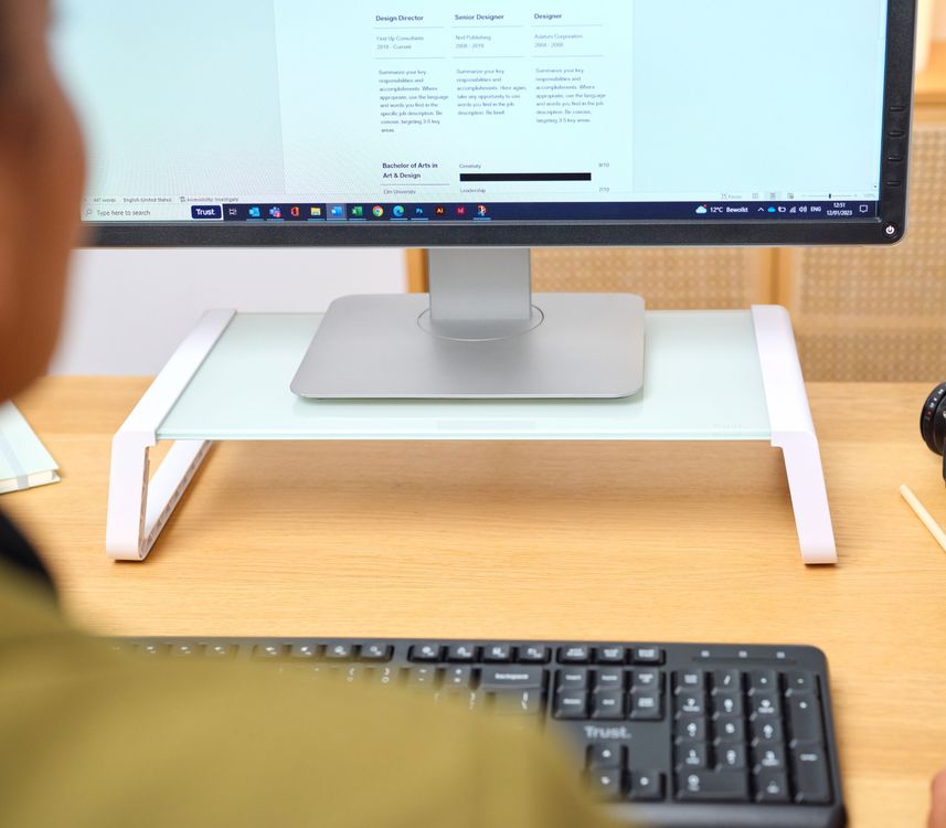 Monitor de computadora sobre un soporte para monitor de cristal templado Trust Monta de color blanco, con teclado en primer plano, sobre un escritorio de madera frente a una persona.